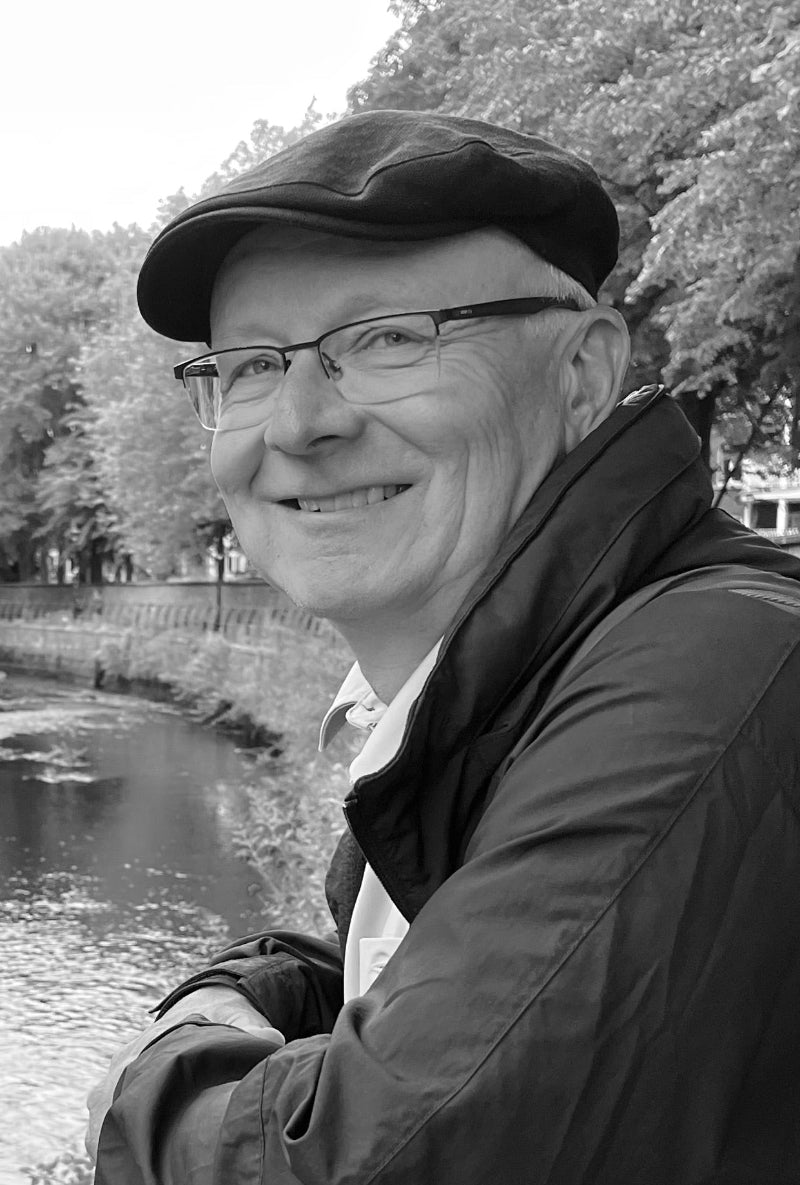 Black and white photo of Stephen Ashton with glasses and a cap by a river.