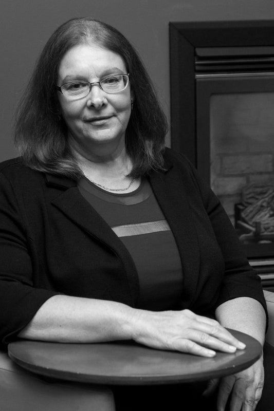 Black and white portrait of a woman sitting at a table with a fireplace in the background.