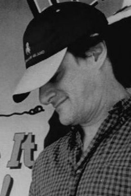 Black and white photo of a person wearing a cap with a logo, sitting in front of a sign.