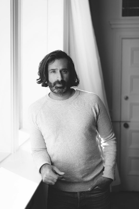 Man wearing a light sweater standing in a room with a window and door in the background.