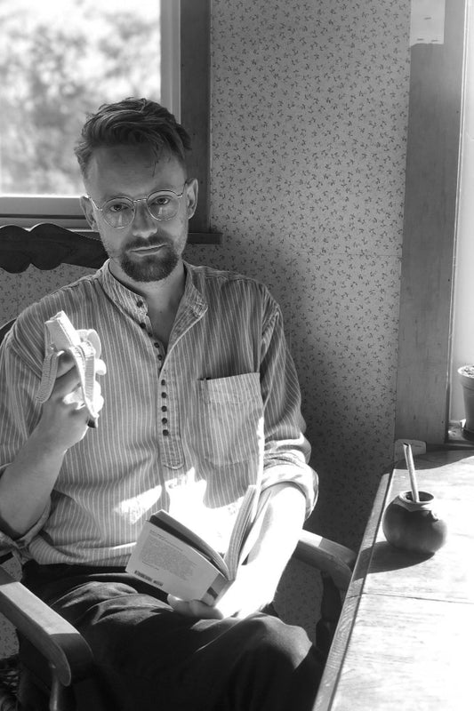 Man sitting on a chair holding a book and a small object, with a window and patterned wall in the background.