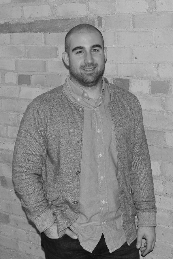 Man wearing a textured sweater over a button-up shirt against a brick wall.