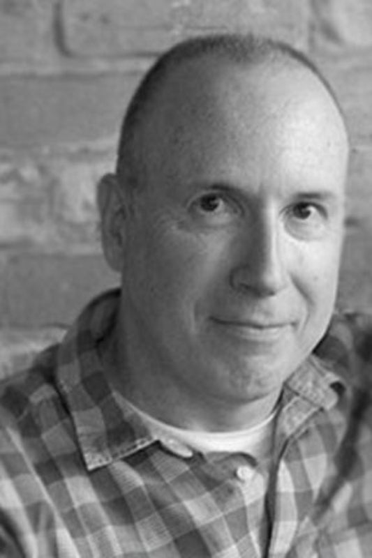Black and white photo of a person wearing a plaid shirt against a brick wall.